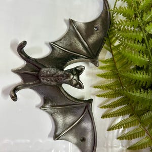 May include: Two metal bat wall decorations with a dark gray finish. The bats have detailed wings and a textured body. The image also includes green fern leaves in the background.