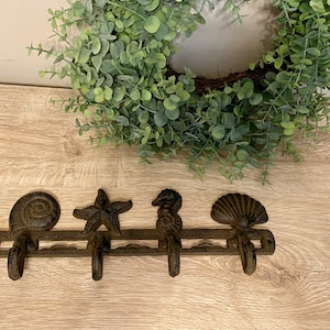 Może przedstawiać: Dekoracyjny, żeliwny wieszak ścienny z czterema haczykami, każdy ozdobiony innym stworzeniem morskim: ślimakiem, rozgwiazdą, konikiem morskim i muszlą. Haczyki są brązowe i zamontowane na poziomej listwie. W tle zielony wieniec z eukaliptusa.