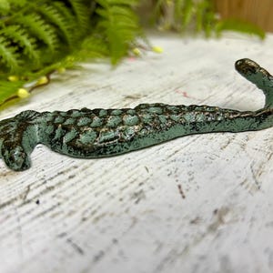 Może przedstawiać: Żeliwny haczyk w kształcie konika morskiego o postarzanym wyglądzie. Konik morski jest pomalowany na turkusowo-zielony kolor z ciemnobrązowymi akcentami, co nadaje mu rustykalny, vintage wygląd. Haczyk jest przeznaczony do montażu na ścianie do wieszania przedmiotów. Ma około 20 cm długości.