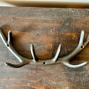 Może przedstawiać: Czarny metalowy hak w kształcie poroża jelenia z pięcioma haczykami. Hak jest zamontowany na drewnianej powierzchni.