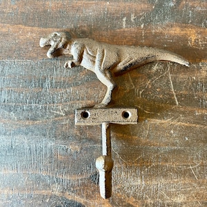 Może przedstawiać: Hak w kształcie dinozaura wykonany z żeliwa, z brązowym wykończeniem. Dinozaur stoi z otwartą paszczą. Hak jest przymocowany do prostokątnej podstawy z dwoma otworami.