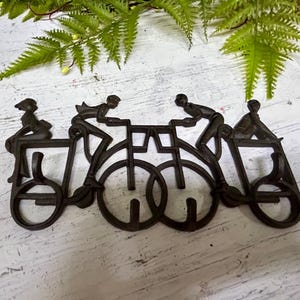 Może przedstawiać: Czarna metalowa dekoracja ścienna przedstawiająca trzy postacie jadące na rowerach. Postacie są stylizowane, a rowery są połączone ze sobą.