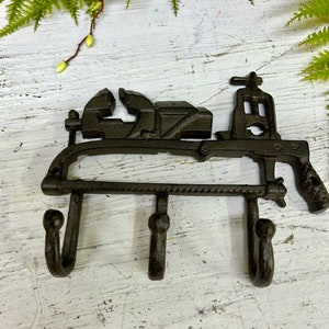 Może przedstawiać: Hak ścienny z żeliwa z trzema haczykami w kształcie imadła, klucza i rury. Hak jest pomalowany na czarno i ma rustykalny wygląd.