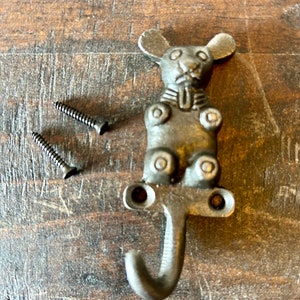 Może przedstawiać: Hak w kształcie myszy wykonany z żeliwa z hakiem w kształcie litery J i dwoma śrubami. Mysz siedzi z ogonem owiniętym wokół haka.