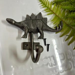 Może przedstawiać: Dekoracyjny wieszak ścienny z motywem dinozaura Stegozaura w ciemnoszarym wykończeniu. Wieszak wykonany jest z metalu i zawiera dwie śruby do montażu. Dinozaur ma szczegółową fakturę i zakrzywiony haczyk do wieszania przedmiotów.