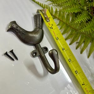 Może przedstawiać: Brązowy metalowy wieszak ścienny w kształcie koguta, z dwiema czarnymi śrubami. Hak ma około 7,6 cm długości, mierzone żółtą miarką. Ozdobna paproć i biała powierzchnia w tle.