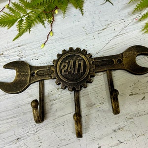 Może przedstawiać: Brązowy metalowy wieszak ścienny w kształcie klucza zębatego z kołem zębatym pośrodku. Na kole zębatym widnieje napis "24/7". Pod kluczem znajdują się trzy haczyki. W tle dekoracyjne gałęzie paproci.