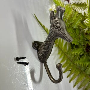 Może przedstawiać: Dekoracyjny metalowy wieszak ścienny w kształcie głowy i szyi żyrafy. Haczyk jest w kolorze brązu z szczegółową fakturą i zawiera dwie czarne śruby do montażu. Głowa żyrafy skierowana jest w prawo.