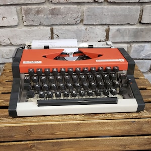 May include: A vintage Olympia typewriter in red and white with black keys. The typewriter is sitting on a wooden crate.