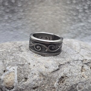 May include: A silver-colored ring with an engraved, repeating scroll design. The ring is wide and has a slightly textured appearance. The ring is sitting on a gray rock.
