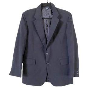 May include: A dark navy blue blazer with subtle vertical stripes. The blazer has a two-button closure, notched lapels, and two front pockets. The sleeves have four buttons at the cuffs. The blazer is hanging on a black hanger.