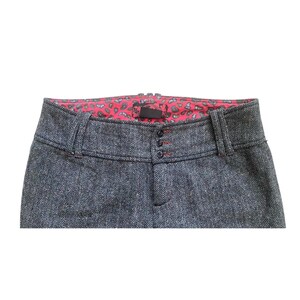 Op de afbeelding: Close-up van een grijze broek met visgraatpatroon. De broek heeft een rode voering met paisley-print zichtbaar bij de taille. Kenmerken zijn een knoopsluiting en riemlussen. De stiksels zijn rood.