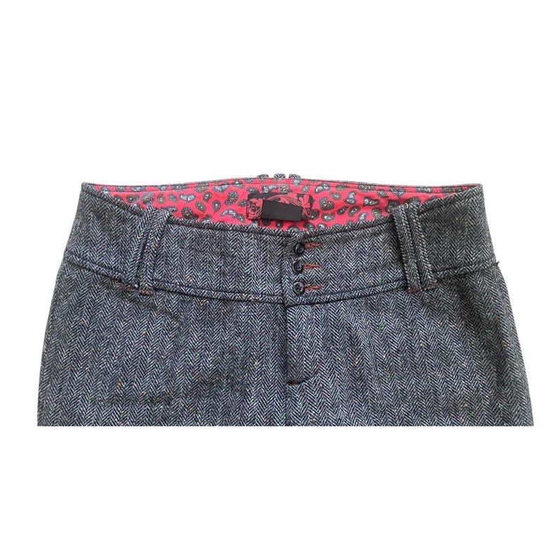 Op de afbeelding: Close-up van een grijze broek met visgraatpatroon. De broek heeft een rode voering met paisley-print zichtbaar bij de taille. Kenmerken zijn een knoopsluiting en riemlussen. De stiksels zijn rood.