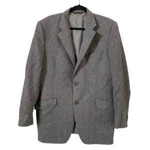 Peut inclure: Un blazer gris avec une fermeture à deux boutons, des revers crantés et deux poches avant. Le blazer est suspendu à un cintre noir. Le tissu du blazer est texturé.