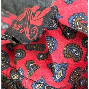 Op de afbeelding: Close-up van een rode stof met een paisley patroon in blauw, bruin en goud. Een zwart label met rood bloemmotief en het woord "Purple" is zichtbaar. De stof is gestikt op een grijs materiaal.