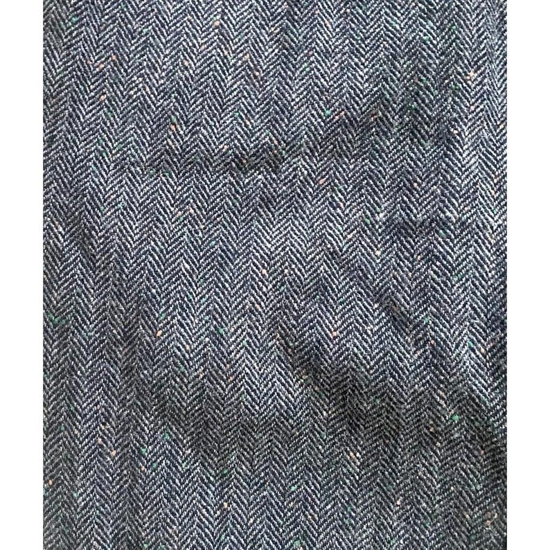 Op de afbeelding: Close-up van een textiel met een visgraatpatroon. De stof is voornamelijk donkerblauw met witte en kleine groene en bruine vlekken. De weving cre&euml;ert een subtiel, diagonaal gestreept effect.