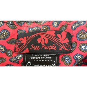 Op de afbeelding: Close-up van een rode paisley stof met een zwart label met de tekst "Free People". Onder het label staat een kleiner label met het nummer "6" en de zin "have a nice day."