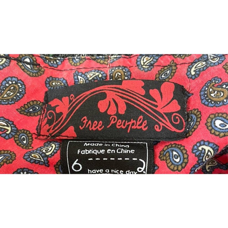 Op de afbeelding: Close-up van een rode paisley stof met een zwart label met de tekst "Free People". Onder het label staat een kleiner label met het nummer "6" en de zin "have a nice day."