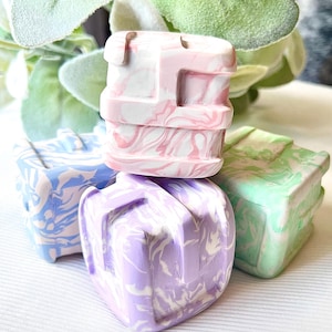 May include: A stack of four small, square-shaped decorative objects with a marbled design. The top object is pink and white, with others in blue, purple, and green. The objects have a geometric design.