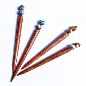 May include: Four wooden pencils with a tapered, reddish-brown grain finish. Each pencil is topped with two decorative, spherical beads featuring a marbled pattern of blue, white, red, and brown colors. The pencils have standard graphite tips.