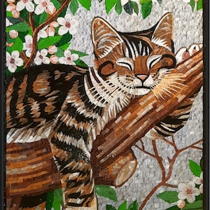 Puede incluir: Una obra de arte en mosaico que representa a un gato atigrado durmiendo en una rama de árbol con flores blancas floreciendo en las ramas de arriba. Los ojos del gato están cerrados y su pelaje es marrón y negro.