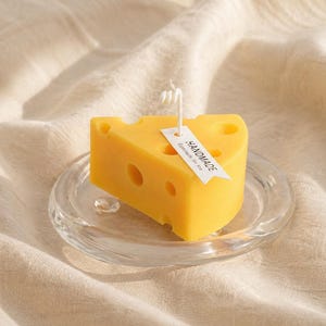 Könnte beinhalten: Eine gelbe, käseförmige Kerze mit Löchern, auf einem transparenten Glasuntersetzer. Ein weißes Etikett mit der Aufschrift "HANDMADE" ist angebracht. Die Kerze hat einen weißen Docht und steht auf einem beigen Stoff.