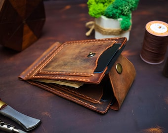 May include: A handcrafted brown leather wallet with a money clip, open to reveal cards and cash. The wallet has a distressed finish and is stitched with a contrasting thread. A small brass button secures the wallet. The background includes tools and decorative items.