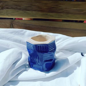 Puede incluir: Pequeña maceta de cerámica azul con un diseño geométrico. La maceta tiene un acabado brillante y un interior beige. Se encuentra sobre una tela blanca, con un fondo de madera.