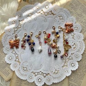 Puede incluir: Una colección de siete pulseras de charms doradas con cuentas de varios colores, charms y un lazo. Los charms incluyen una mariposa, una campana, una flor y una estrella. Las pulseras se muestran sobre un encaje blanco.