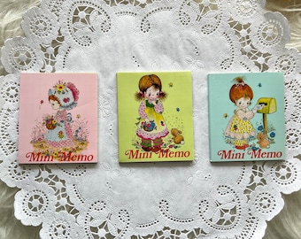 May include: Three square Mini Memo notepads with illustrations of little girls. The notepads are pink, green, and blue. Each notepad has the words "Mini Memo" in red. The notepads are on a white lace doily.