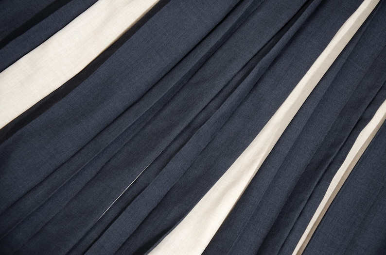 May include: Diagonal view of a fabric with alternating navy blue and off-white stripes. The fabric appears to be pleated or folded, creating a textured surface with shadows and highlights.