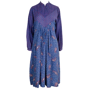 Puede incluir: Un vestido azul de manga larga con cuello alto. El corpiño presenta un diseño estampado en rojo y azul, mientras que la falda tiene un estampado de cachemira. El vestido tiene un diseño fluido y plisado.
