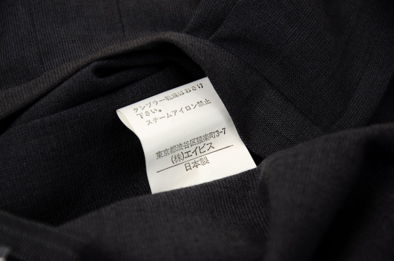 May include: Close-up of a black fabric with a white tag attached. The tag has Japanese text and the words "Made in Japan". The fabric is folded, showing its texture and the tag's details.
