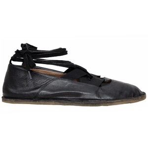 May include: A black leather flat shoe featuring a rounded toe, a low profile, and black fabric laces that crisscross over the top of the foot and tie around the ankle. The shoe has a thin, flat, light-coloured sole and a smooth, flexible construction.