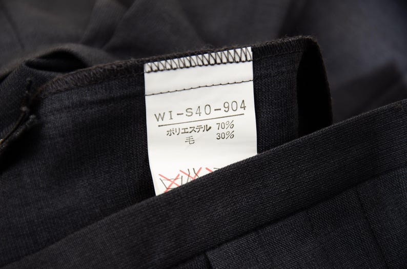 May include: Close-up of a black garment with a white tag attached. The tag displays the text "WI-S40-904" and Japanese characters indicating the fabric composition: 70% polyester and 30% wool. The garment's fabric appears to be a dark, solid color.