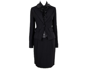 May include: A black skirt suit with a matching blazer and a ruffled neckline. The blazer has a single-button closure and two front pockets. The skirt is knee-length. The suit is made of a solid black fabric.