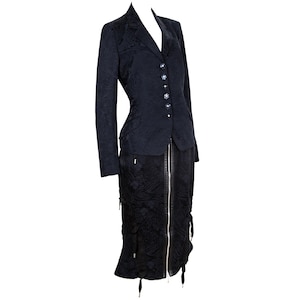 May include: A dark navy blue suit consisting of a blazer and a skirt. The blazer has a notched lapel, decorative buttons, and long sleeves. The skirt has a front zipper and decorative ties.