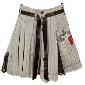 May include: A light gray pinstriped skirt featuring a dark brown ribbon waistband and front tie closure. The skirt includes a button-up front, a frayed hemline, and a patch pocket on the right side decorated with red floral embroidery and beads.