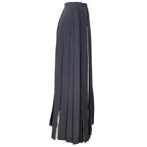 May include: A dark gray pleated skirt with a high waist and a long, flowing design. The skirt has a slit on the side, revealing a lighter-colored fabric underneath. The skirt is made of a textured material.