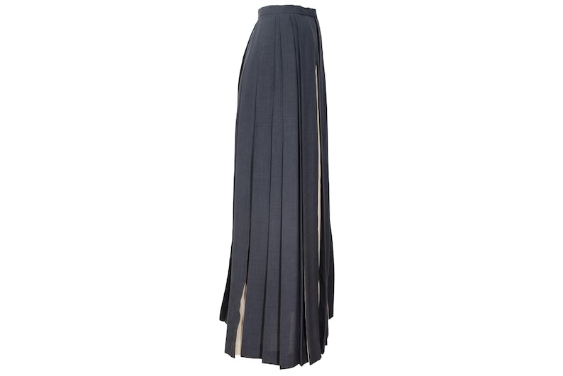 May include: A dark gray pleated skirt with a high waist and a long, flowing design. The skirt has a slit on the side, revealing a lighter-colored fabric underneath. The skirt is made of a textured material.