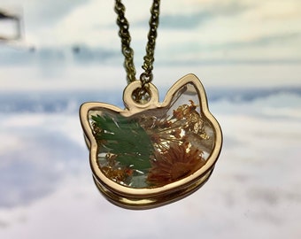 May include: Gold chain necklace with a clear resin pendant shaped like a cat's head. The pendant contains pressed flowers and leaves in shades of green and orange.
