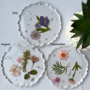May include: Three clear resin coasters with pressed flowers and silver glitter. The coasters are labeled "July", "August", and "September".