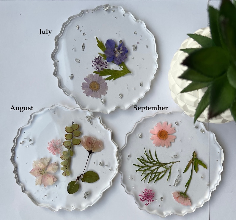 May include: Three clear resin coasters with pressed flowers and silver glitter. The coasters are labeled "July", "August", and "September".