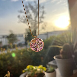 May include: A gold chain necklace with a round pendant featuring a clear resin filled with dried flowers and leaves. The pendant is a light pink and purple color.