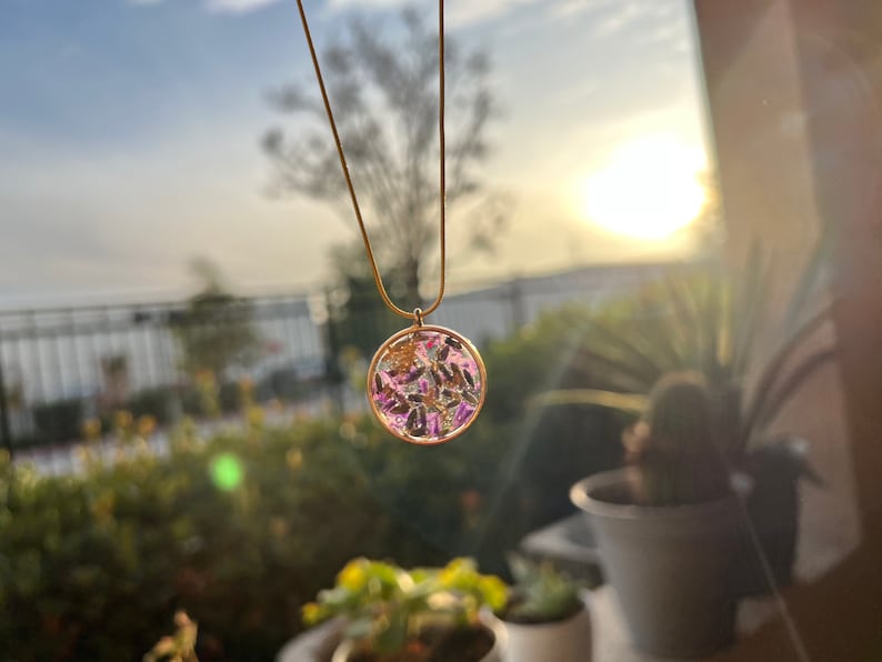 May include: A gold chain necklace with a round pendant featuring a clear resin filled with dried flowers and leaves. The pendant is a light pink and purple color.