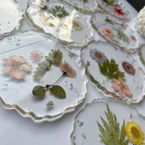 May include: A set of clear resin coasters with pressed flowers and leaves. The coasters have a wavy edge and are decorated with various dried flowers and leaves, including white, pink, yellow, and green.