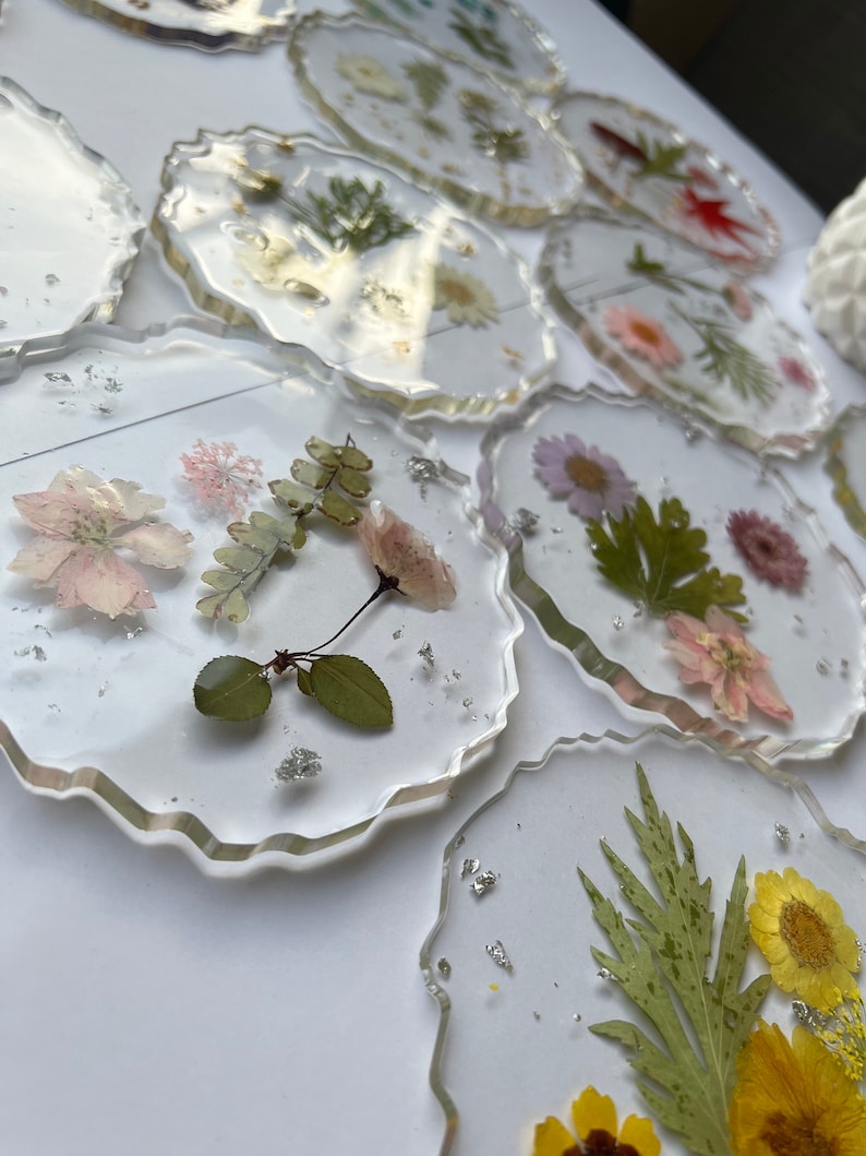 May include: A set of clear resin coasters with pressed flowers and leaves. The coasters have a wavy edge and are decorated with various dried flowers and leaves, including white, pink, yellow, and green.