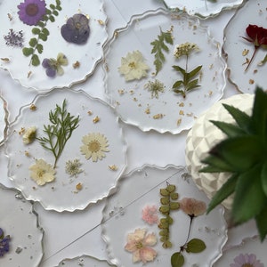 May include: Set of eight round clear resin coasters with gold flakes and pressed dried flowers and leaves. The coasters feature a variety of flowers and leaves in shades of purple, white, pink, and green.