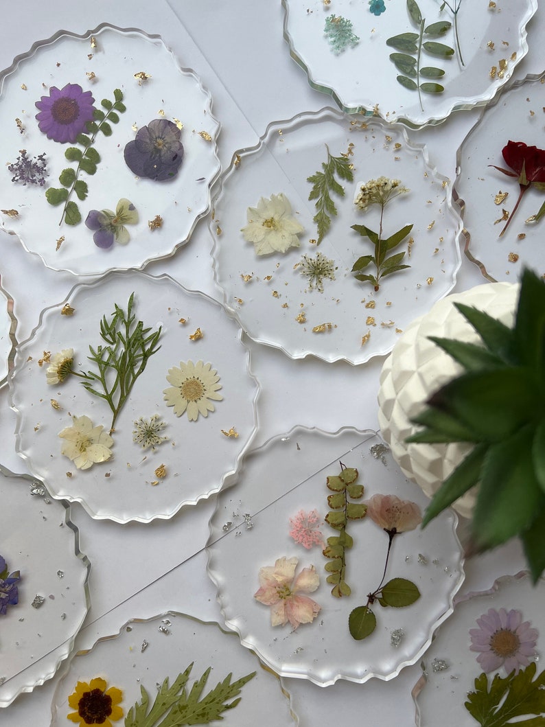 May include: Set of eight round clear resin coasters with gold flakes and pressed dried flowers and leaves. The coasters feature a variety of flowers and leaves in shades of purple, white, pink, and green.