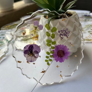 May include: A clear resin coaster with a scalloped edge features pressed purple and yellow flowers, green ferns, and gold flecks. The coaster is sitting on a white surface.